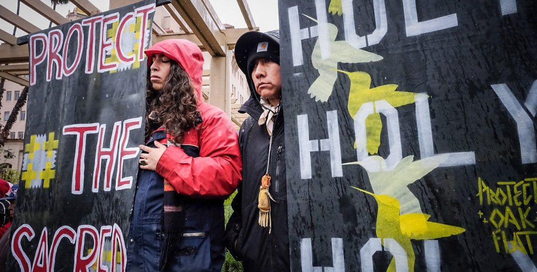 Una pareja de mujeres indígenas racializadas en una protesta pacífica con pancartas defendiendo territorios sagrados.