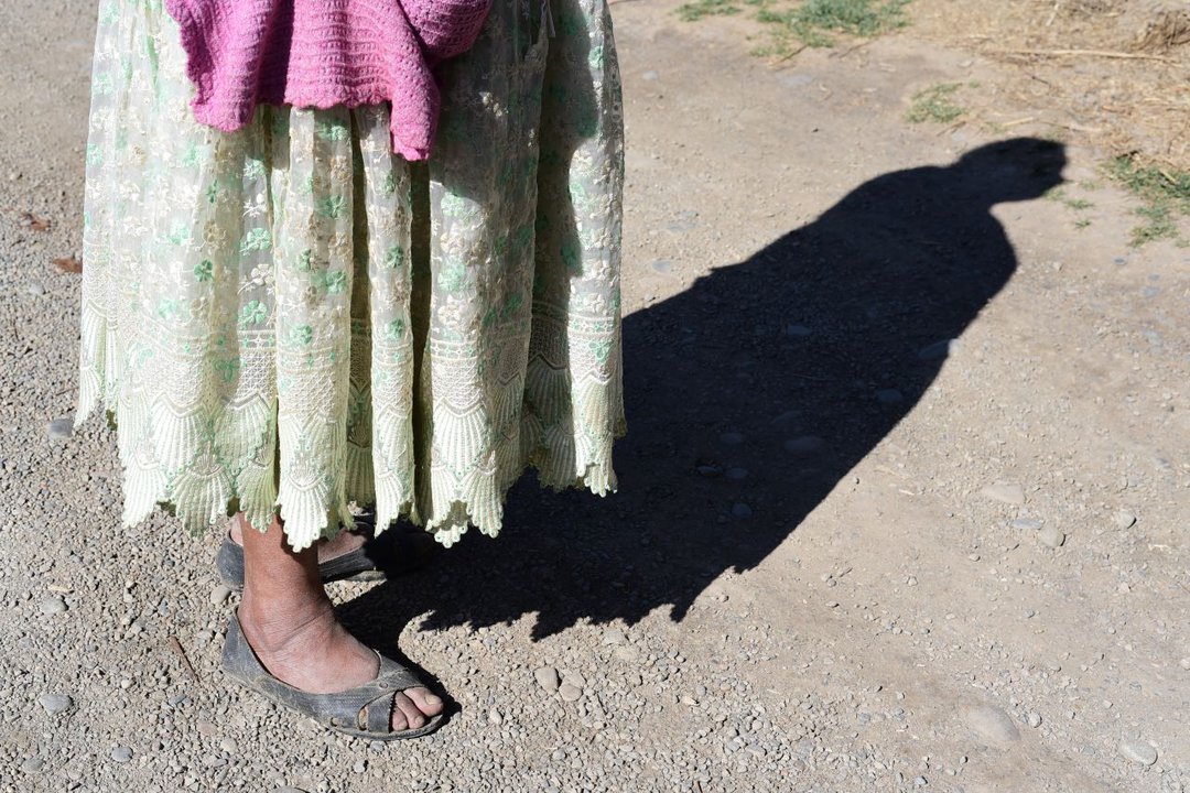 Fotografía de los pies de una mujer agricultora en Mecapaca, La Paz. La sombra de su torso se proyecta hacia la derecha.
