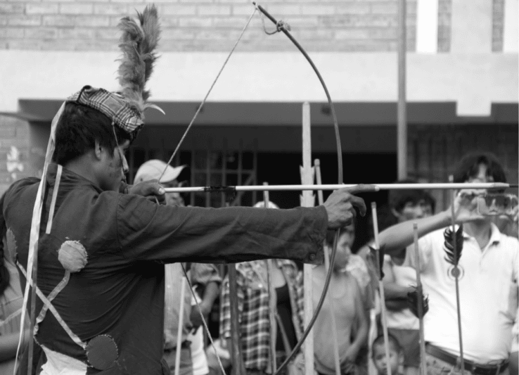 Un joven indígena mosetén sostiene un arco y apunta con él, posando para la fotografía. La fotografía está en blanco y negro y tiene gran simetría.
