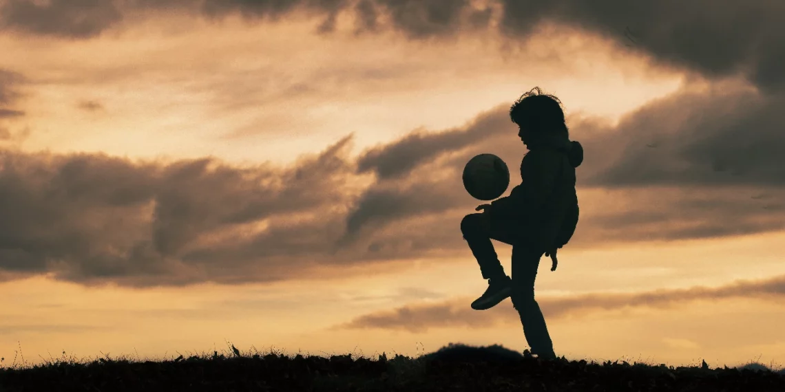 Un niño juega con una pelota de fútbol a contra luz en un paisaje con césped.