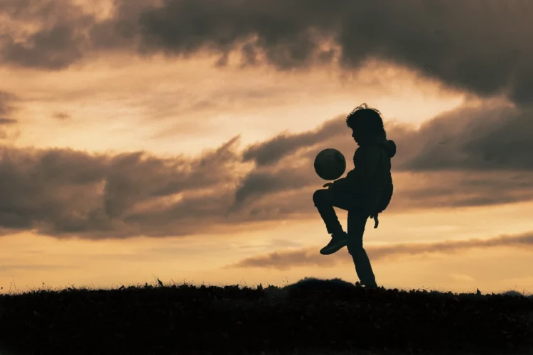 Un niño juega con una pelota de fútbol a contra luz en un paisaje con césped.