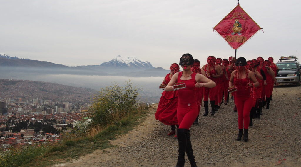 Un fotograma de la película Revolución Puta en la que un grupo de mujeres vestidas de rojo camina por una ladera de la ciudad de La Paz, con el Illimani de fondo.